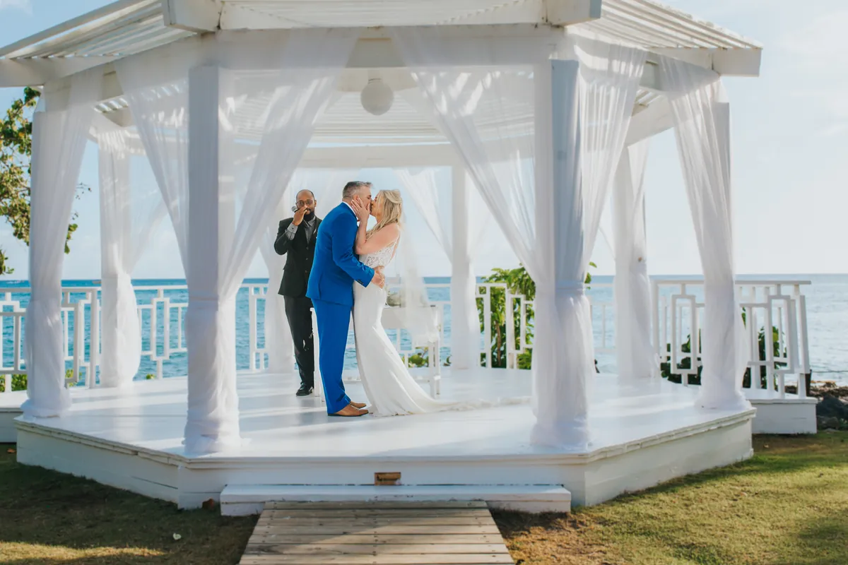 Couple's first kiss in a white gazebo by the sea, officiant behind