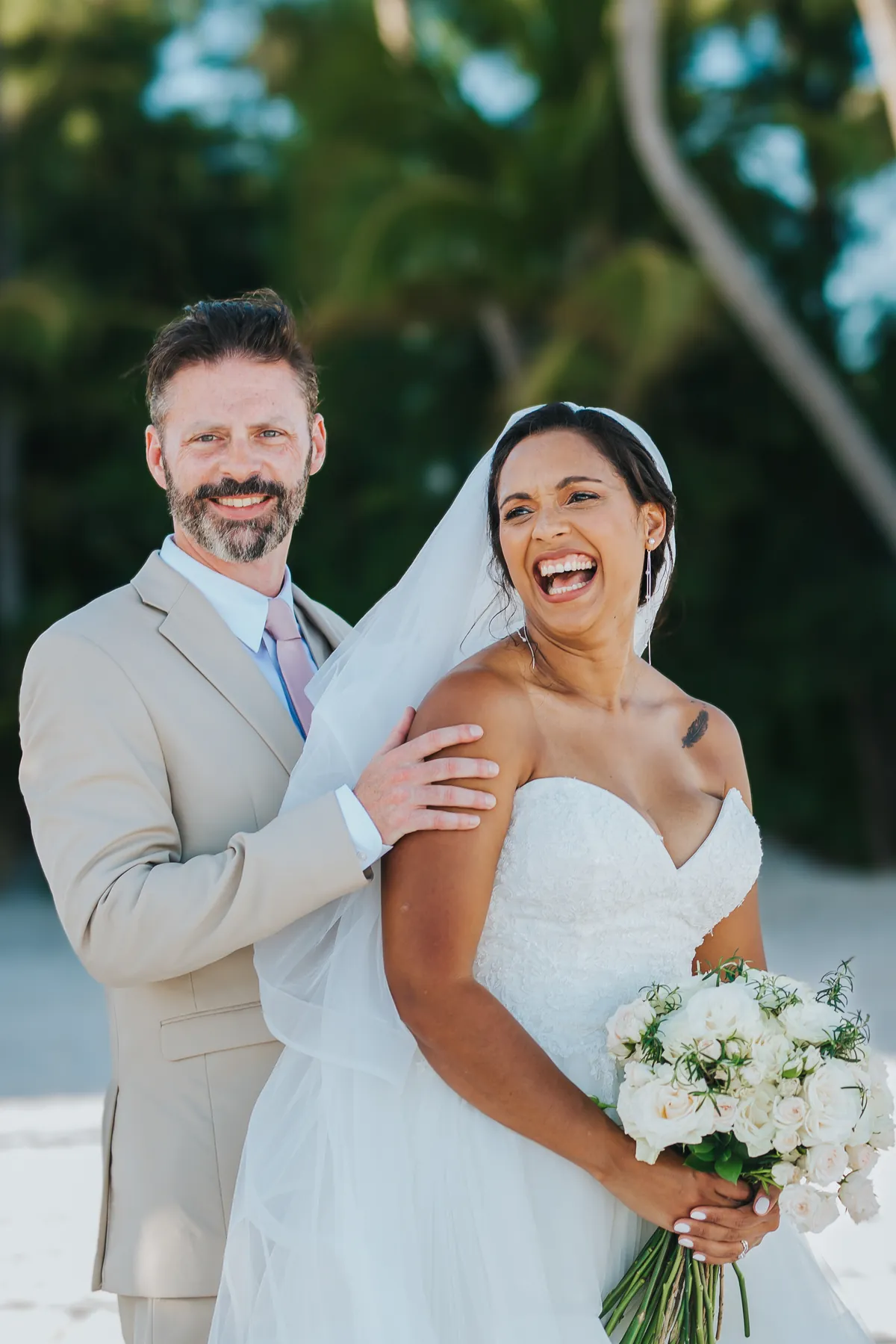 Bride laughing while the groom smiles behind her on the beach